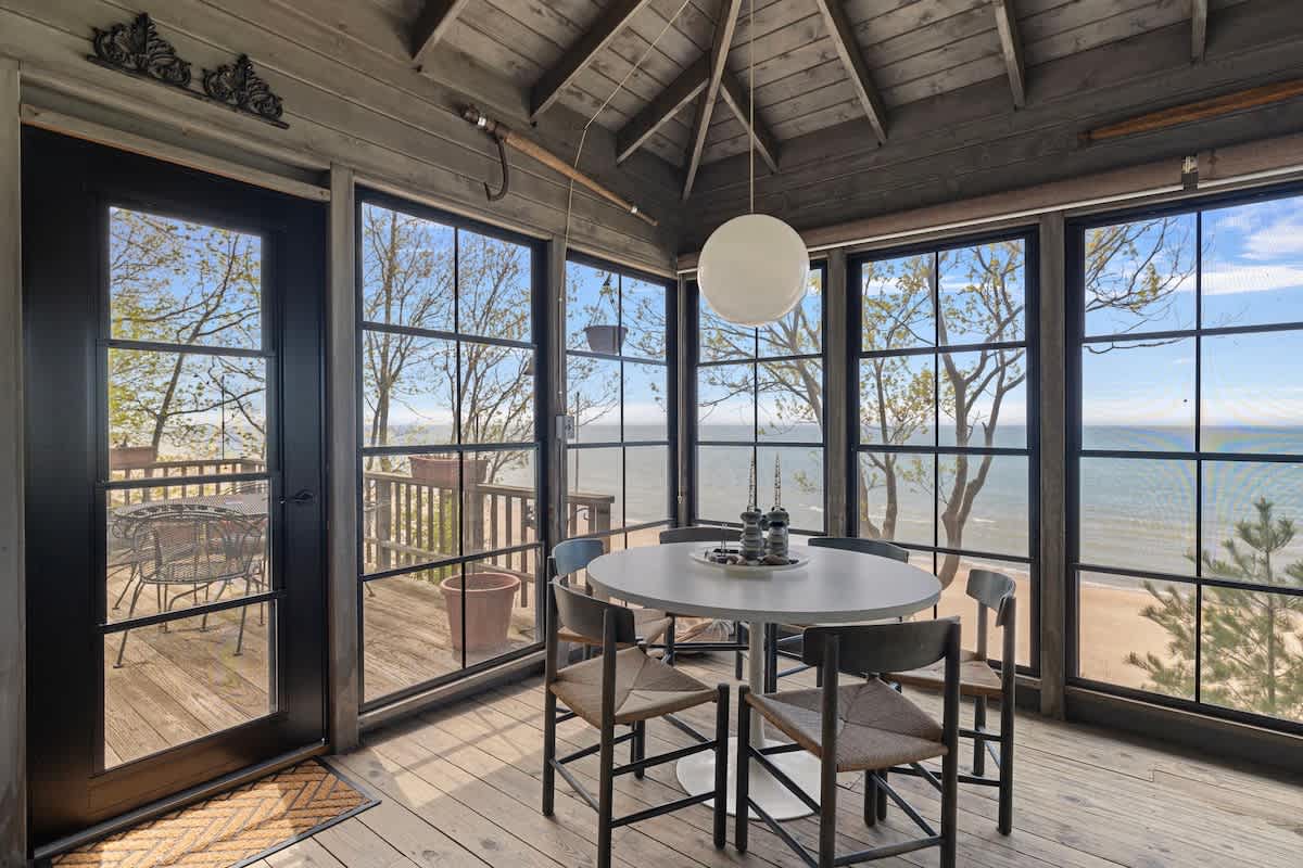 A bright dining area with a round white table and four chairs is surrounded by large windows offering panoramic views of a sandy beach and the ocean. The room has a vaulted wooden ceiling and dark wood floors.
