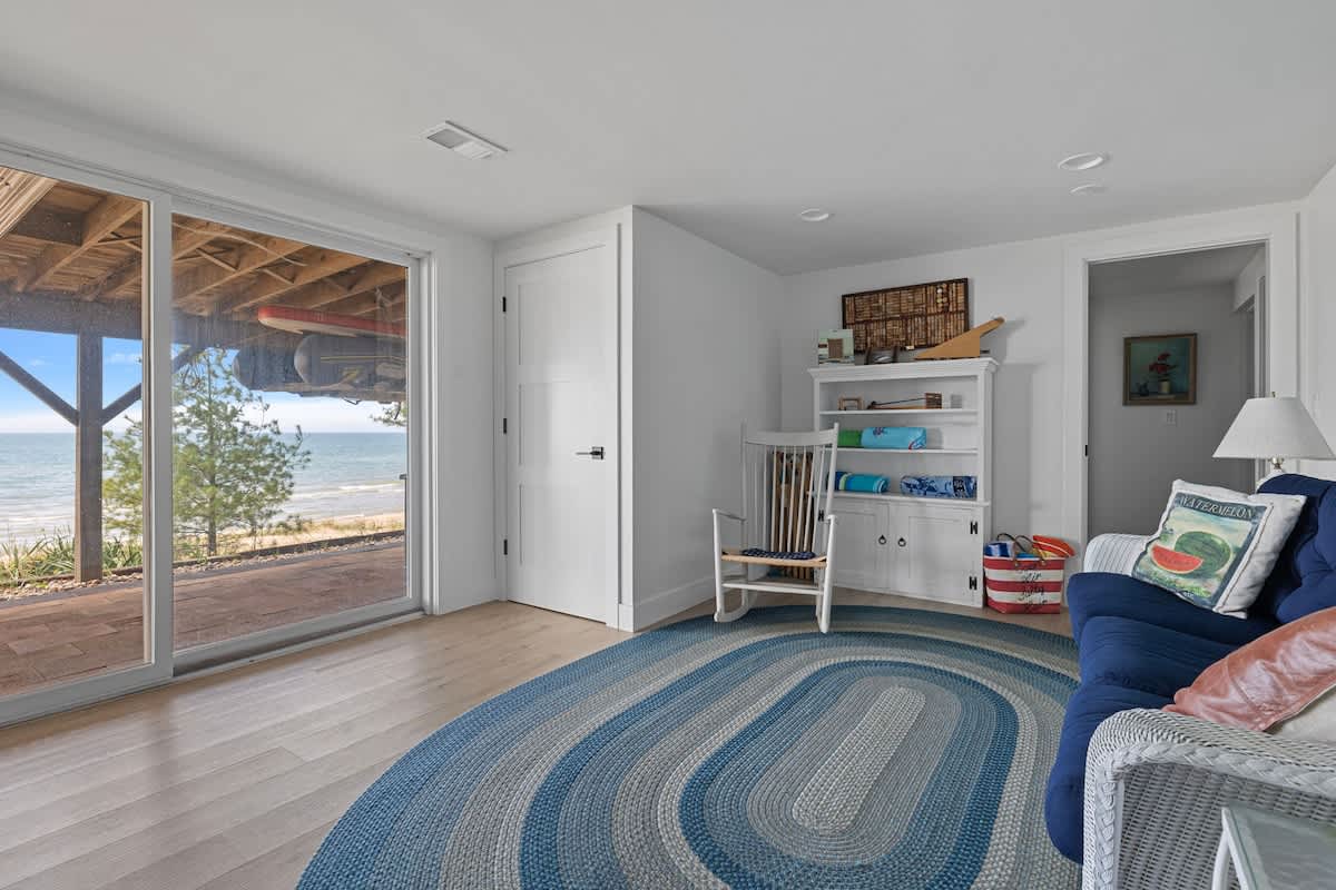 Bright living area with light wood flooring and white walls, featuring a blue braided rug and a blue sofa. Large sliding glass doors open to an outdoor patio with a view of the beach and water.