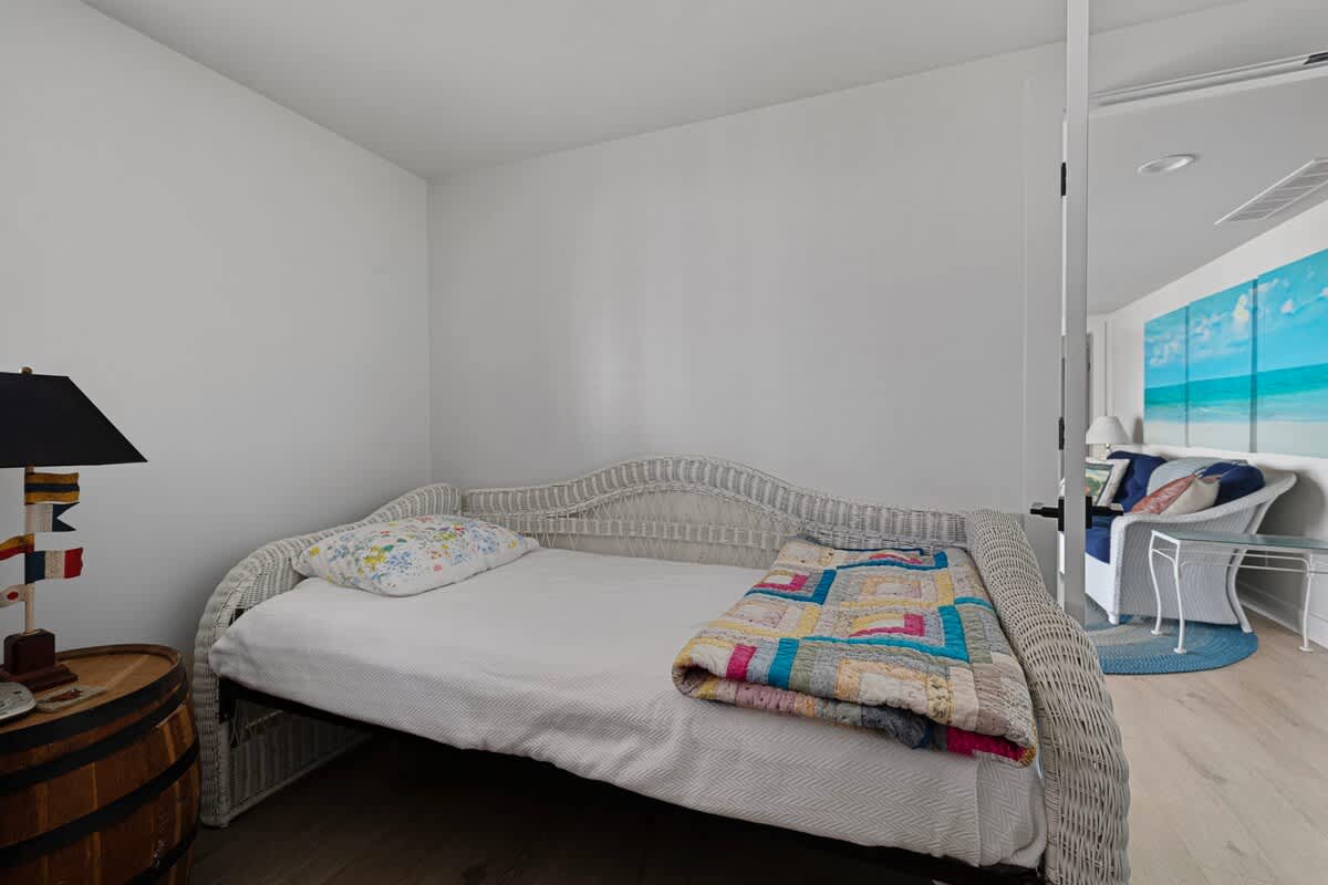 A cozy bedroom with a white wicker daybed adorned with a colorful quilt, a rustic barrel nightstand, and a lamp with a black shade, set against white walls.