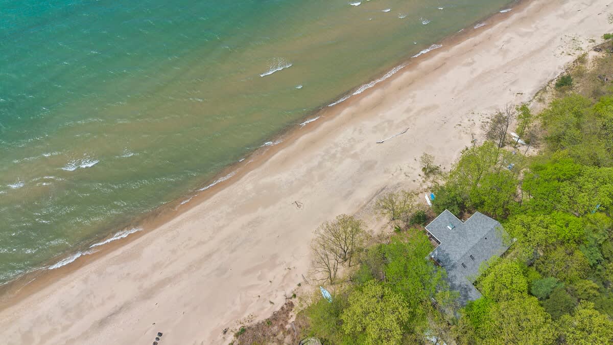 A high aerial shot capturing the vibrant turquoise water meeting a wide sandy beach, with a single house surrounded by dense green foliage.