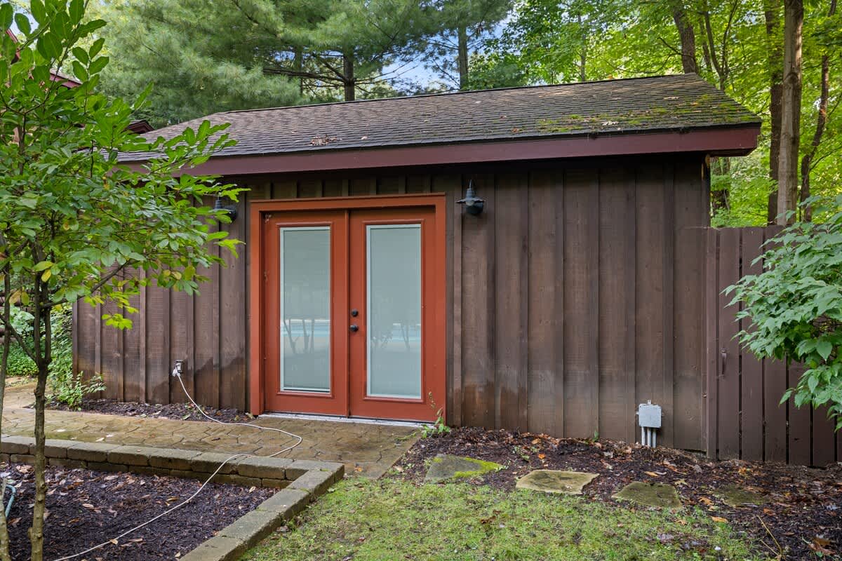 Detached wooden structure with a dark brown exterior and red double doors, set amidst a natural landscape with trees and shrubs. The building features a dark shingled roof and outdoor lighting fixtures above the doors, suggesting a garage or shed.