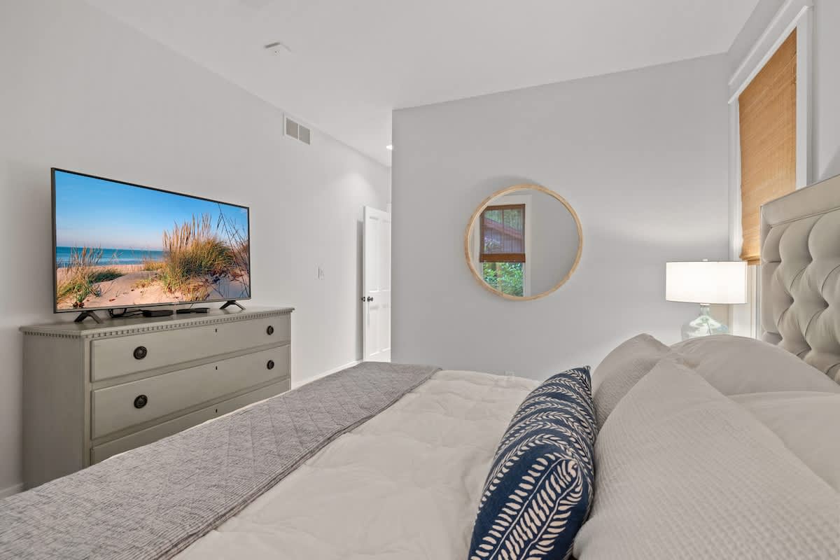 A view from the bed in a bright bedroom, showing a flat-screen TV on a gray dresser, a round mirror on the wall, and a glimpse of a doorway.