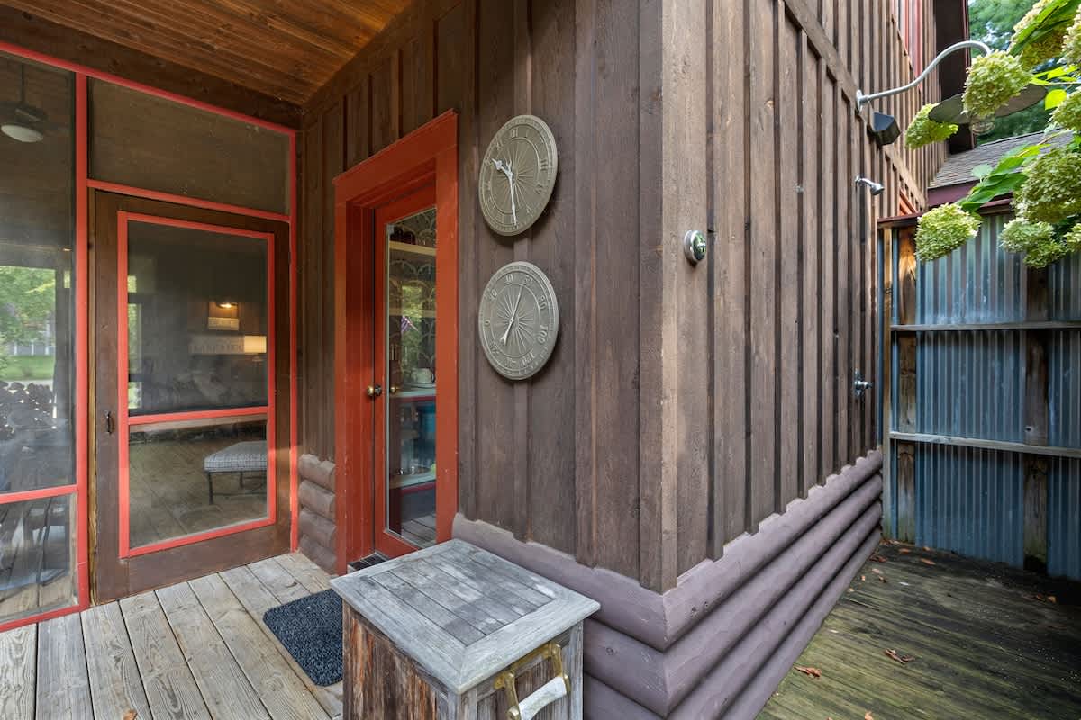 Close-up of a rustic wooden house exterior featuring a screened porch entrance with a vibrant red door and an adjacent outdoor shower with corrugated metal siding. Two decorative wall hangings adorn the dark wood siding, and a wooden storage box sits on the deck.