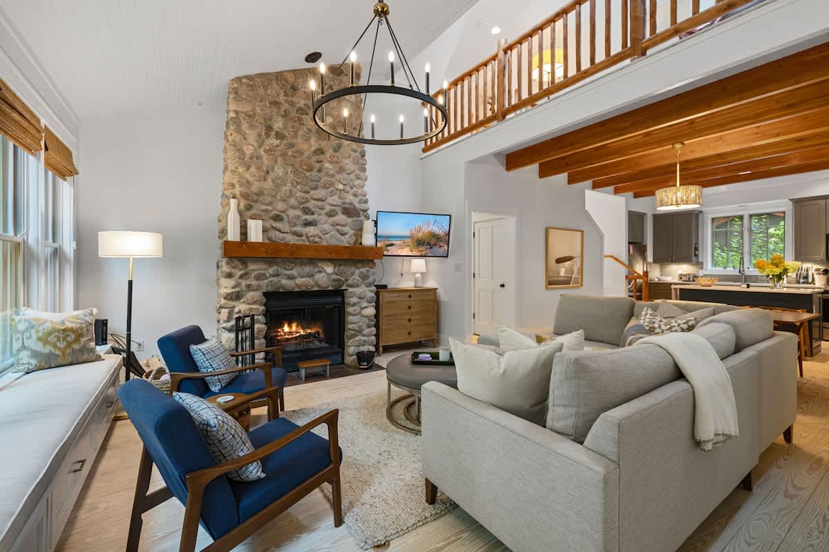 Spacious living room with a towering stone fireplace and a large L-shaped sectional sofa, offering an open view towards the kitchen and an upper-level balcony, all under a high white ceiling with wooden beams.