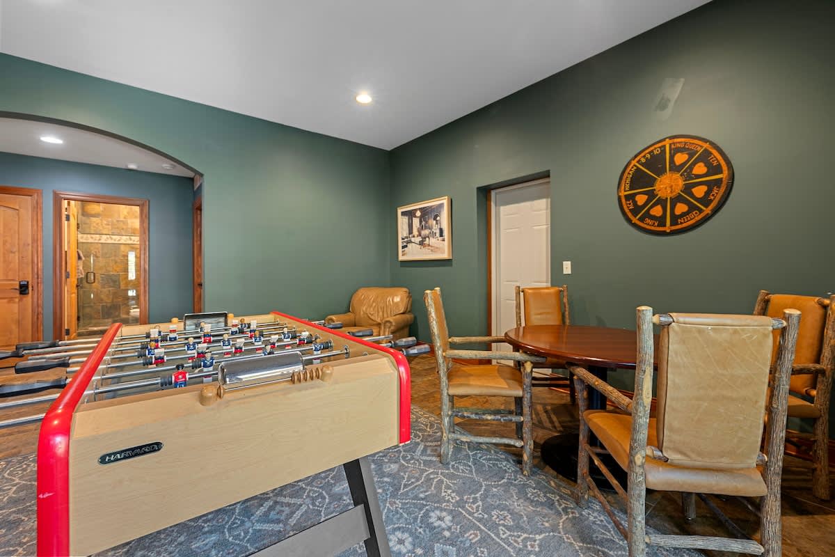 A game room with dark green walls and a patterned area rug, featuring a foosball table and a round wooden table with rustic chairs. A large circular wall art piece is visible.