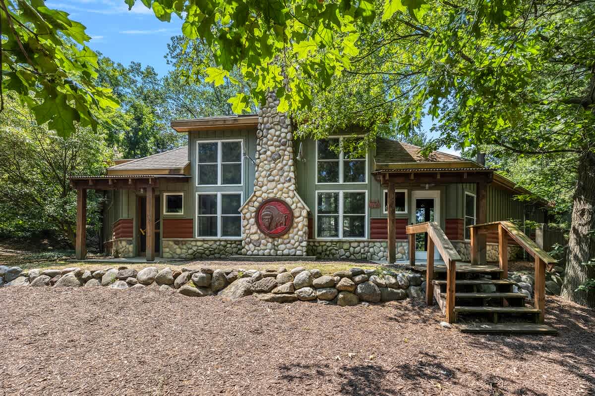 The front exterior of a rustic-style house featuring a prominent stone chimney with a decorative red medallion, large windows, and a wooden porch with steps, nestled among green trees.