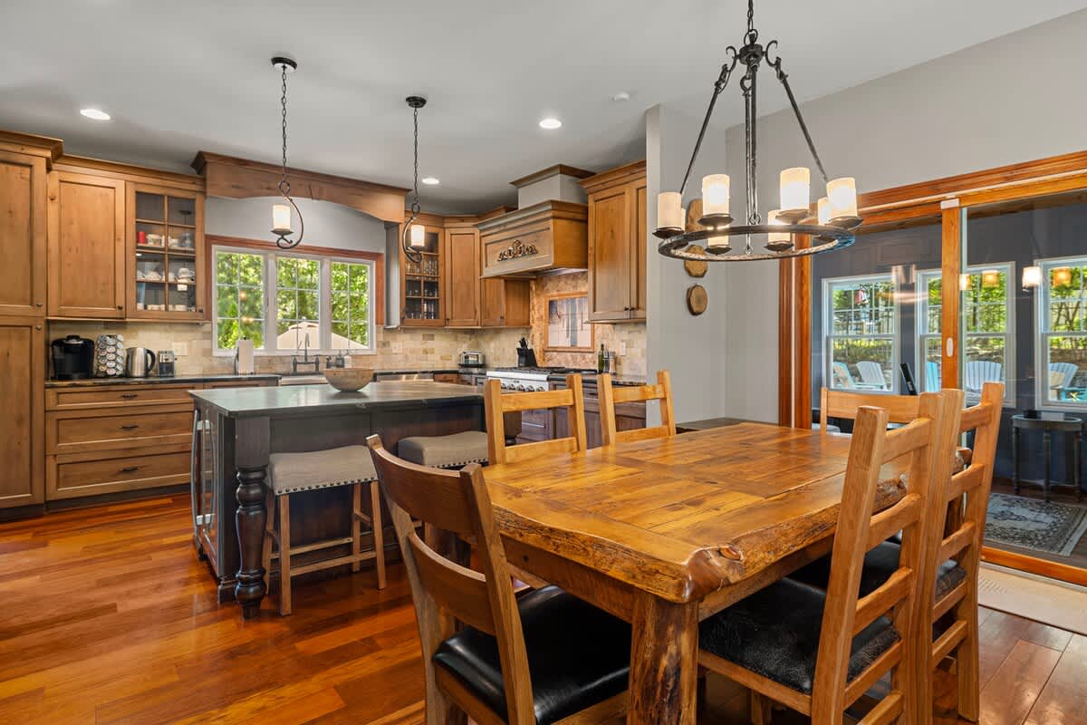 A bright kitchen and dining area showcases warm wooden cabinetry, hardwood floors, and a large wooden dining table with chairs. A kitchen island with a dark countertop is central, and sliding glass doors open to an outdoor space.