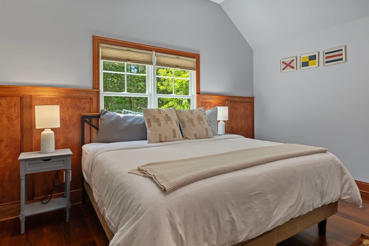 A cozy bedroom features a queen-sized bed with neutral bedding, flanked by two nightstands with lamps. Wooden wainscoting lines the lower walls, and a large window offers views of greenery. Nautical flag art decorates the wall above the bed.