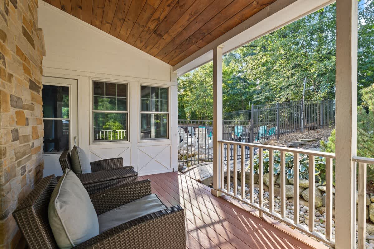 A covered outdoor porch with a rustic stone wall on one side, a wooden ceiling, and white railings, furnished with dark wicker chairs and light cushions, overlooking a fenced patio area.