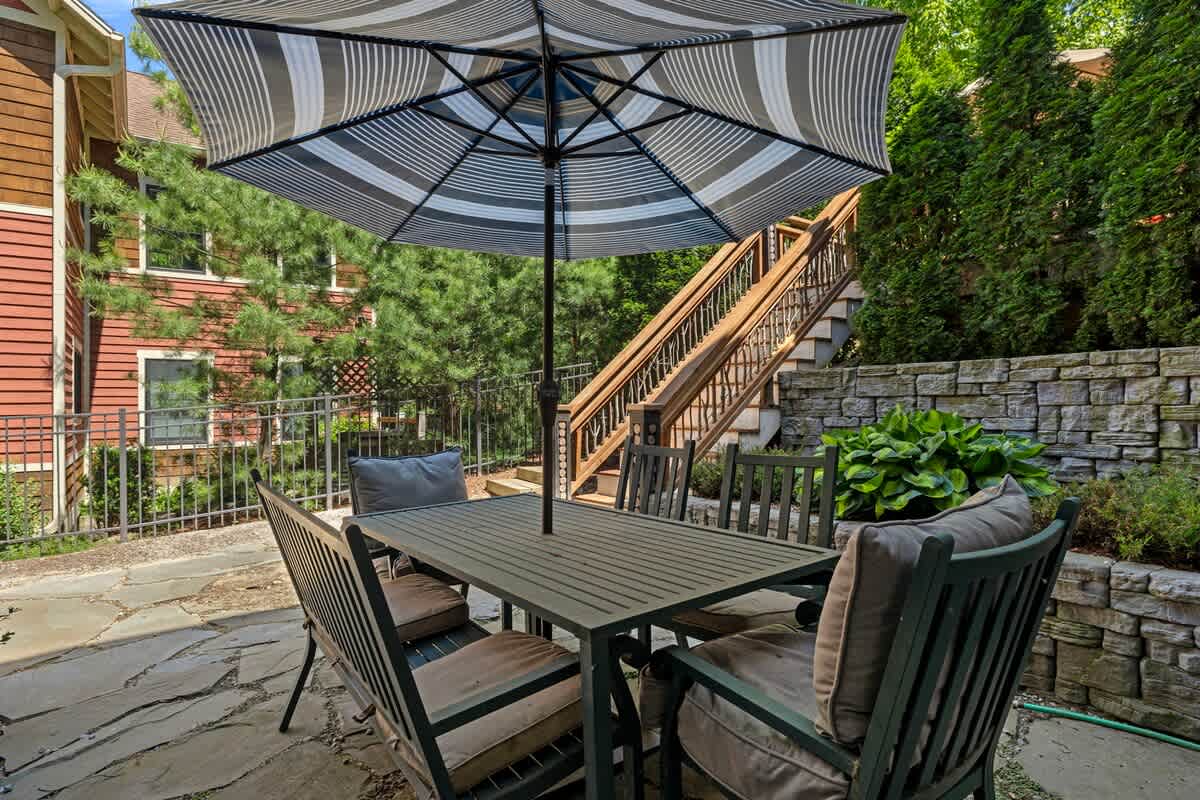 An outdoor stone patio furnished with a rectangular dining table and cushioned chairs under a striped umbrella, bordered by a stone retaining wall and lush greenery, with wooden stairs visible in the background.