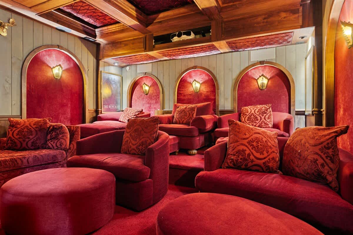 A luxurious home theater room is adorned in rich red velvet, featuring multiple plush armchairs and ottomans arranged in rows. The ceiling has wooden beams and recessed lighting, and arched alcoves with sconces add to the opulent ambiance.