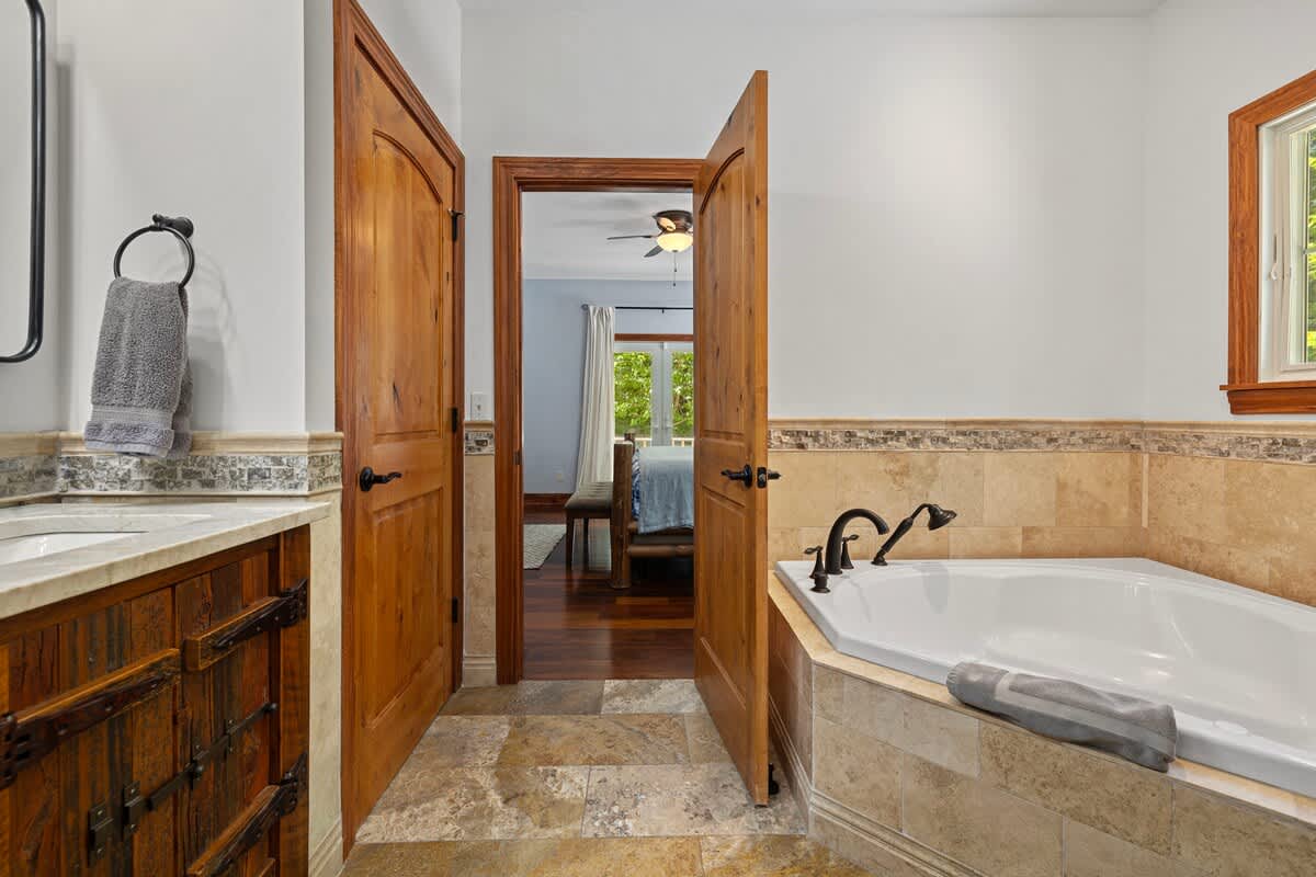 A spacious bathroom with light-colored tiled floors and walls, featuring a large corner jetted tub and a rustic wooden vanity with a sink. Two wooden doors lead to an adjacent bedroom.