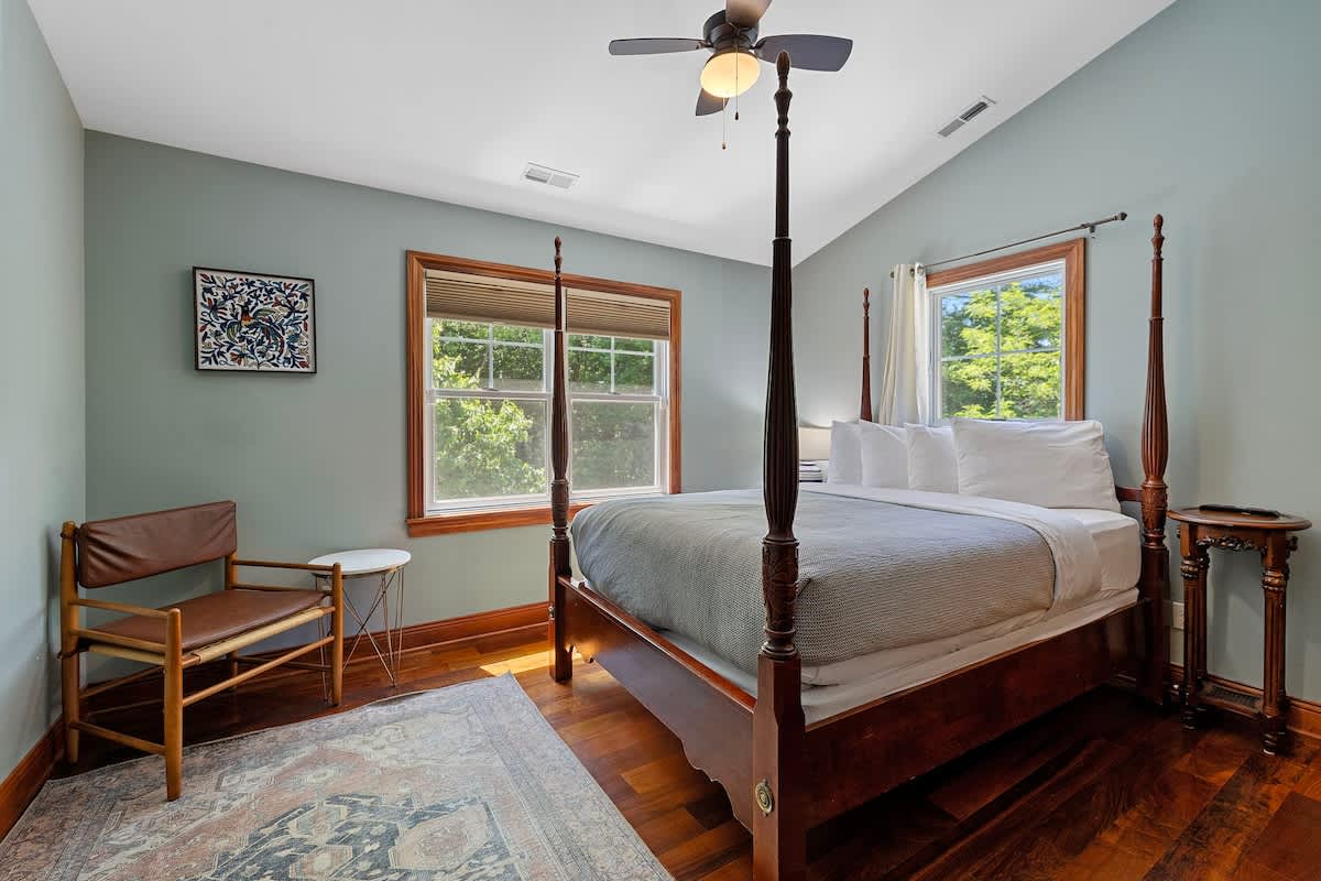 A bright bedroom with light green walls and dark hardwood floors, featuring a dark wood four-poster bed with white bedding. Two large windows offer views of lush greenery, and a ceiling fan is visible.