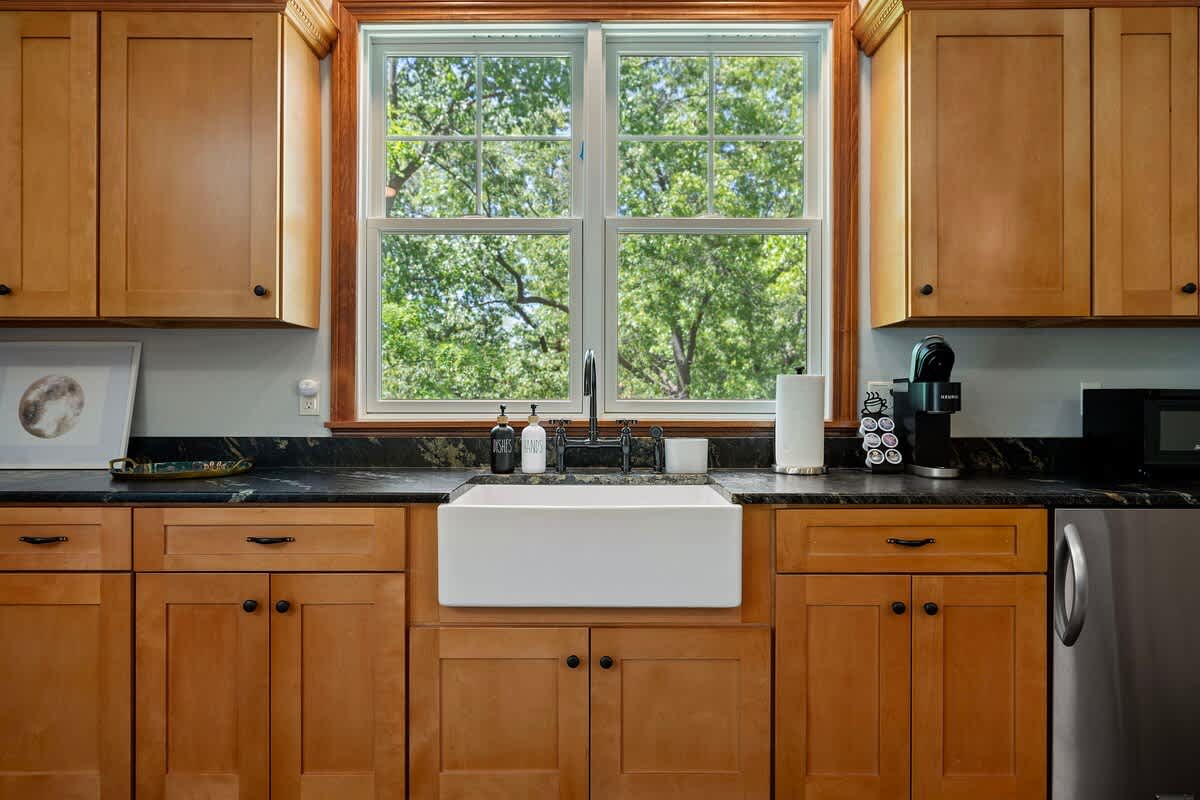 A close-up of a kitchenette area featuring light wood cabinetry, dark countertops, a white farmhouse sink, and a window overlooking lush trees. A coffee maker and microwave are visible.