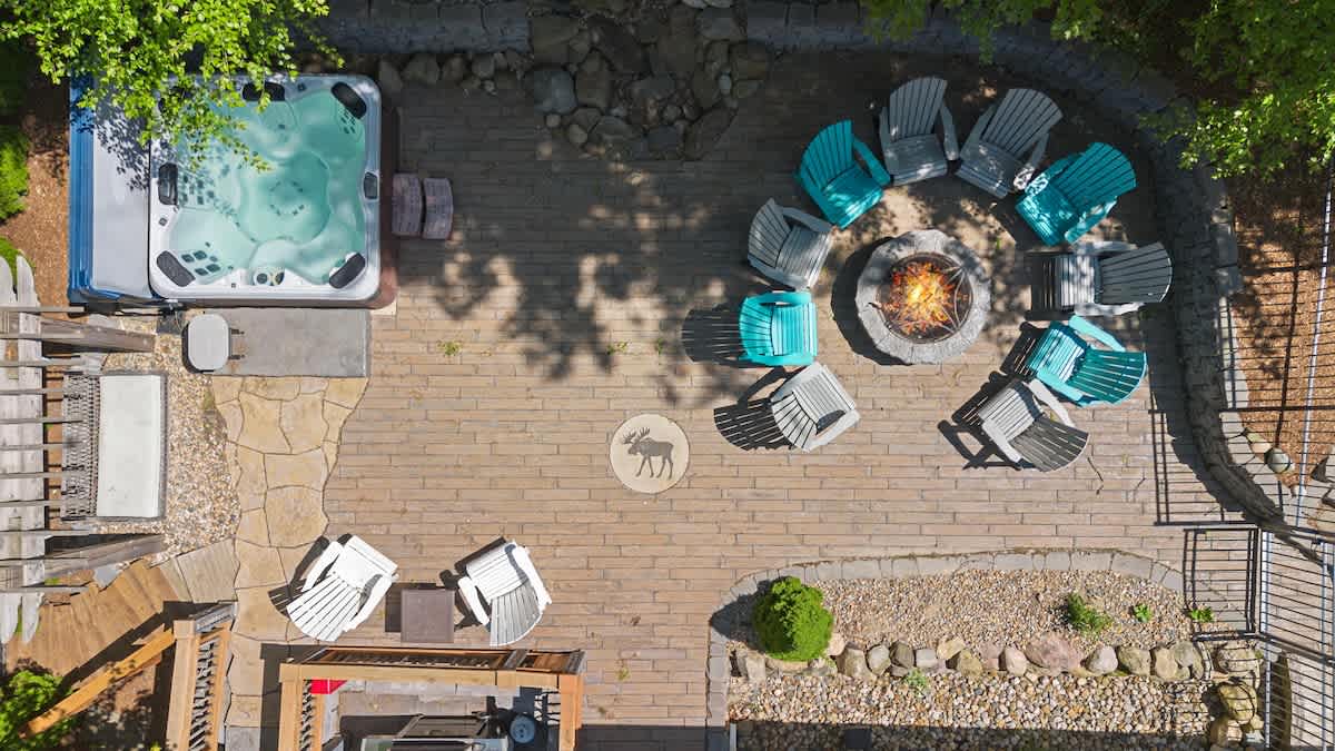 An aerial view of a stone patio featuring a square hot tub, a circular fire pit surrounded by teal and gray Adirondack chairs, and an embedded moose design.