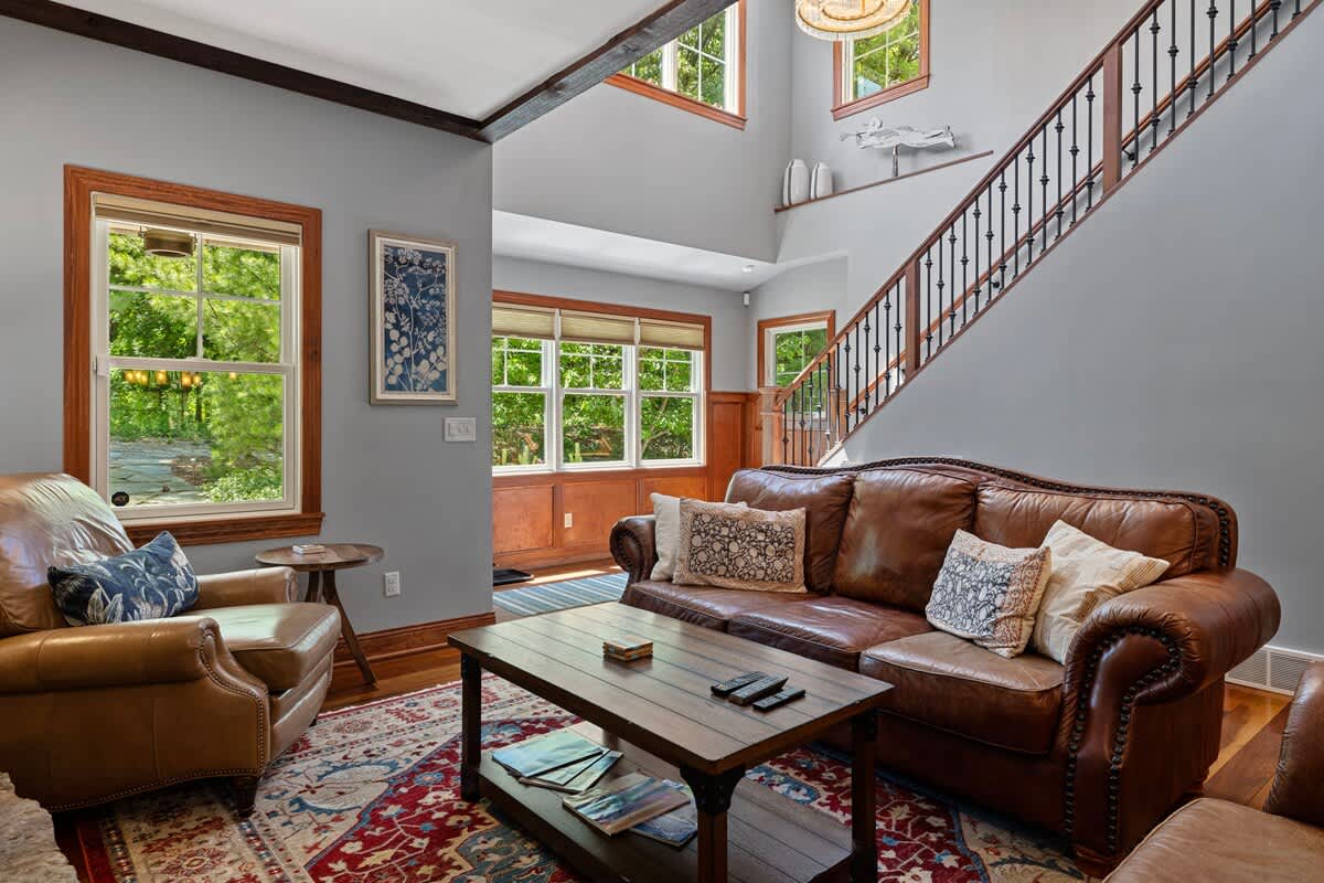 A spacious living room with high ceilings, light gray walls, and a grand staircase with wrought iron balusters, furnished with brown leather seating and a patterned rug.