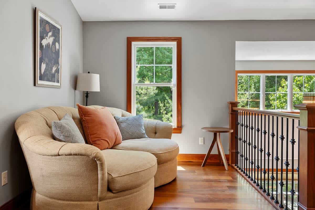 A bright upstairs landing or sitting area with light gray walls, hardwood floors, and a comfortable chaise lounge, overlooking the lower level through a railing.