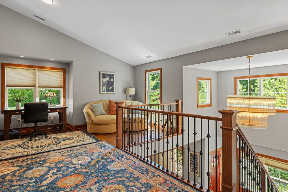 A spacious loft area with light gray walls and hardwood floors features a dedicated workspace with a desk and chair by a window. A cozy armchair and ottoman provide a reading nook, and a decorative rug adds color to the open space overlooking a lower level.
