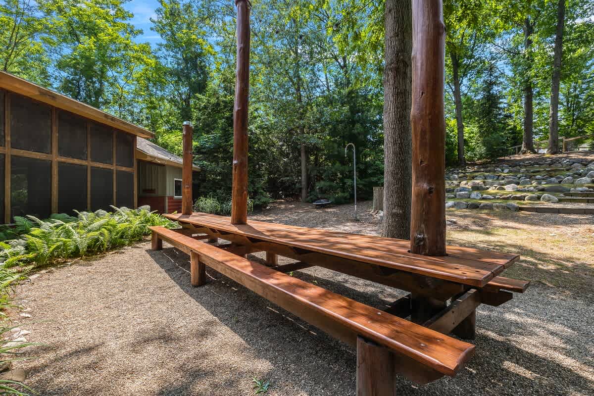 A large, rustic wooden picnic table with benches stands on a gravel patio, surrounded by tall trees and lush greenery, with a screened porch visible on the left side of a building.