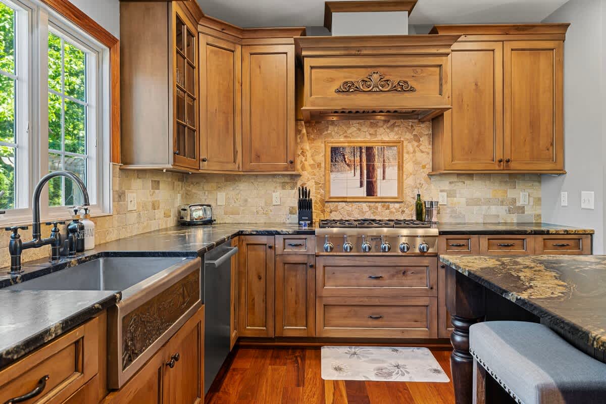 A rustic kitchen featuring custom wooden cabinetry, dark granite countertops, a stone tile backsplash, and a professional-grade stainless steel range.