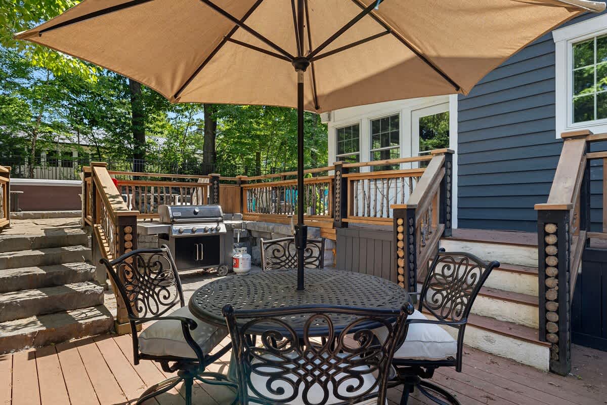 A multi-level wooden deck featuring a round metal dining table with an umbrella and matching chairs, alongside a BBQ grill, with stairs leading to other sections of the outdoor space.