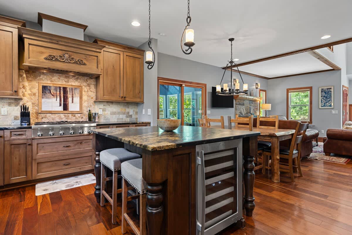 A spacious kitchen with warm wooden cabinetry and hardwood floors features a large island with a dark granite countertop and two bar stools. Stainless steel appliances, including a wine fridge, are integrated, and a dining area with a wooden table and chairs is visible in the background.