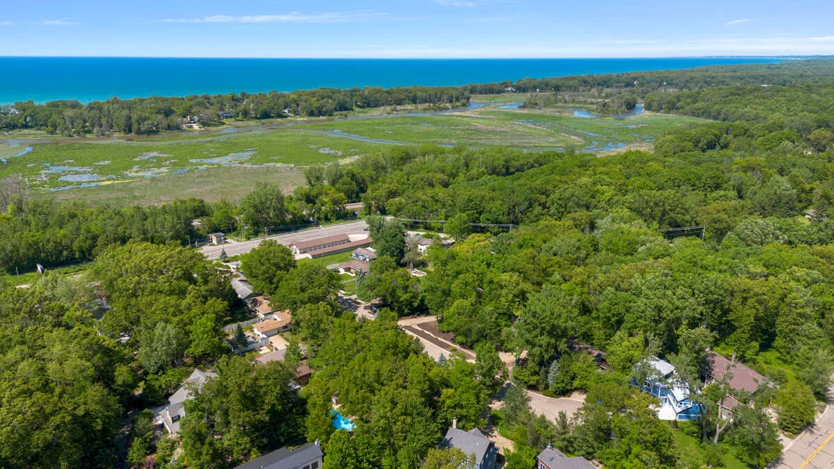 An aerial view showcasing the property nestled within a dense green forest, with a large body of water (likely Lake Michigan) visible in the distance under a clear blue sky, highlighting the natural surroundings.