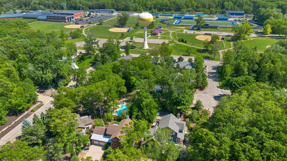 An aerial view of the property and surrounding neighborhood, featuring residential homes amidst lush trees, with a school complex, sports fields, and a water tower visible in the background.