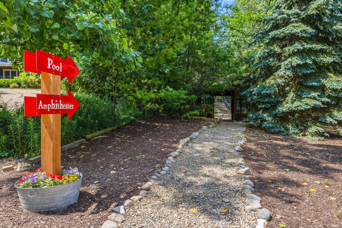 A rustic wooden sign with red arrows pointing towards "Pool" and "Amphitheater," situated beside a gravel path lined with rocks and surrounded by lush trees and a potted flower arrangement.