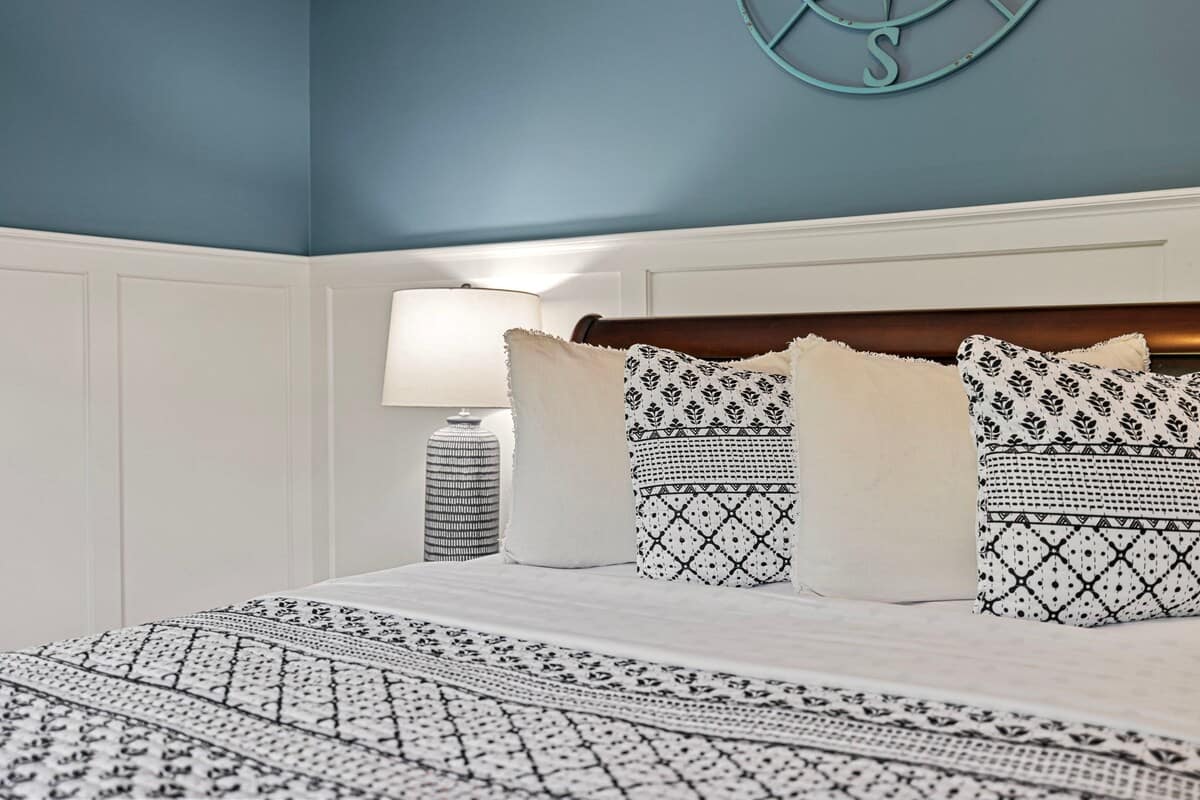 Close-up of a dark wood sleigh bed with white and black patterned bedding and pillows, flanked by a nightstand with a white lamp. The room features blue walls with white wainscoting.
