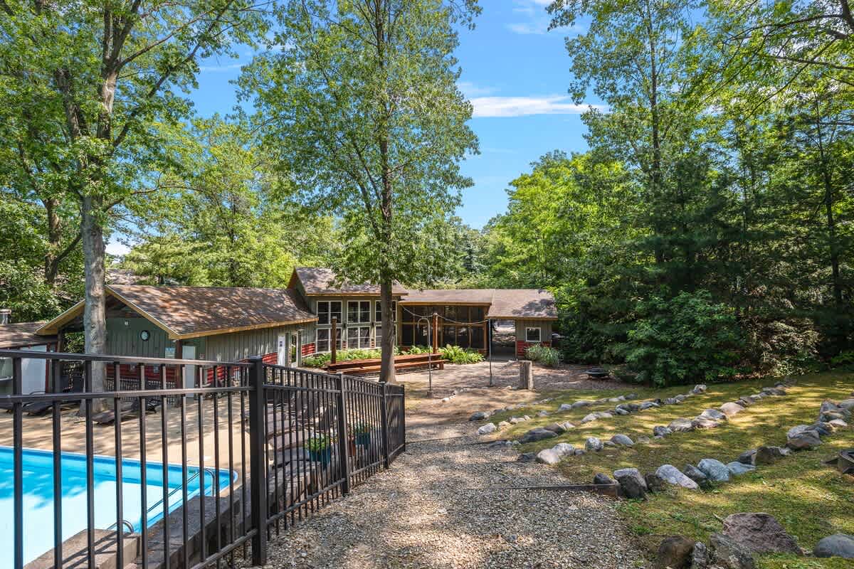 An inviting outdoor swimming pool with a black metal fence in the foreground, surrounded by lush green trees and a rustic-style house with a screened porch in the background under a bright blue sky.
