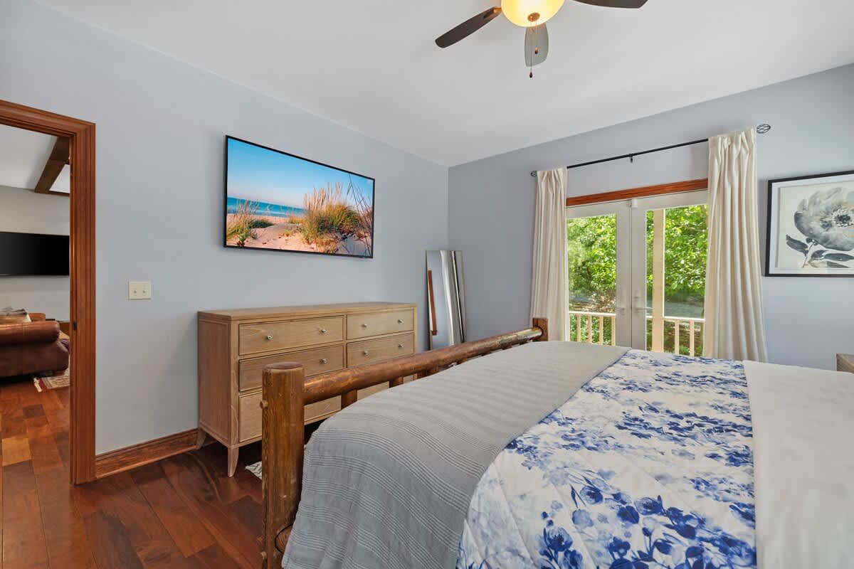 A light blue bedroom features a wooden bed frame with blue and white floral bedding, a wall-mounted TV displaying a beach scene, and a wooden dresser. French doors lead to an outdoor area, and a full-length mirror stands in the corner.