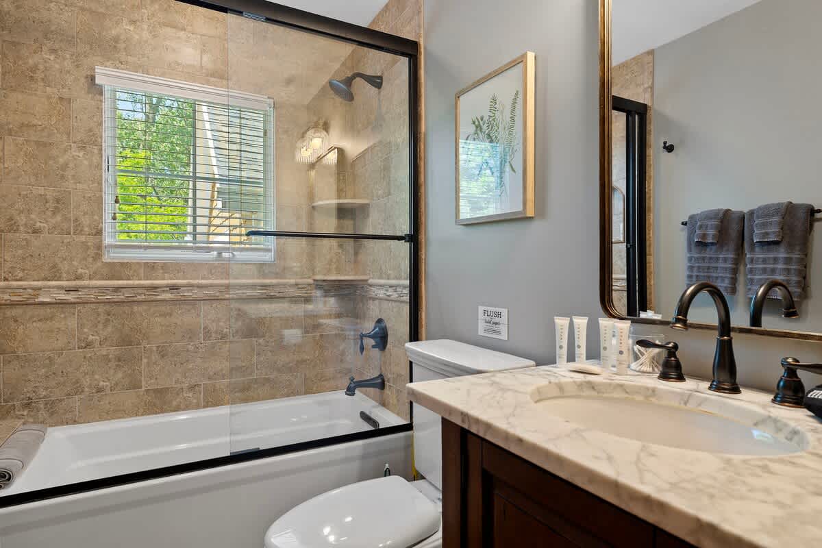 A modern bathroom with light brown tiled walls and a shower/tub combo enclosed by a glass sliding door. A dark wood vanity with a marble countertop and a large mirror completes the space.