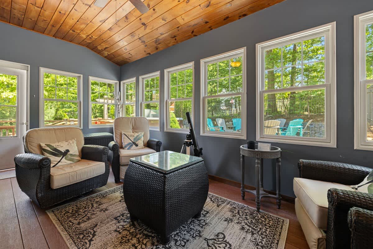 An enclosed sunroom with a vaulted wooden ceiling, large windows offering views of lush greenery, and comfortable wicker armchairs with light cushions arranged around a glass-top coffee table. A telescope stands in the corner.