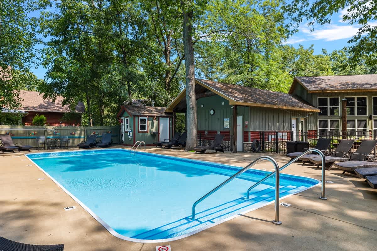 A bright outdoor swimming pool with clear blue water, surrounded by a concrete deck with lounge chairs, and a small pool house visible in the background, all set amidst mature trees.