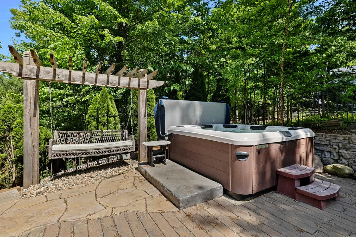 An outdoor hot tub area set on a stone patio, surrounded by lush green trees, featuring a wooden pergola with a swing and steps leading up to the hot tub.