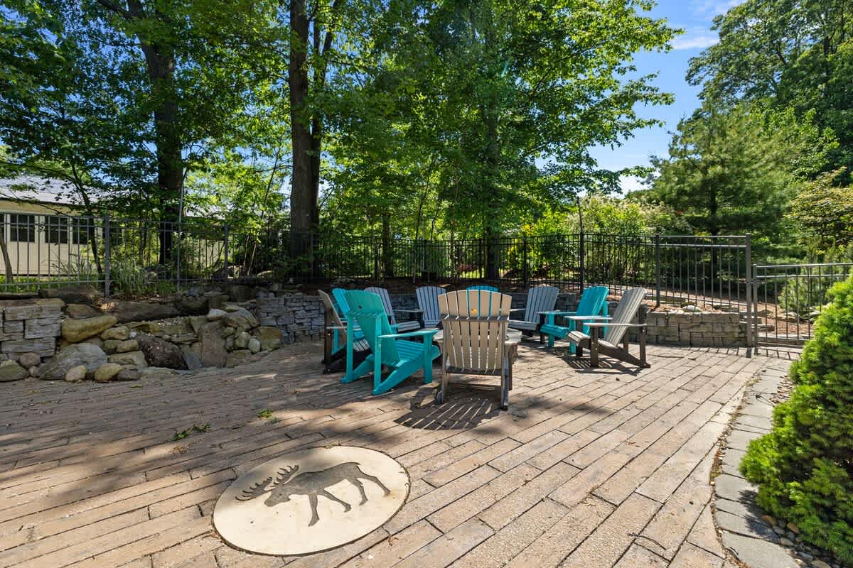 A spacious outdoor patio paved with brick pavers, featuring a circular arrangement of colorful Adirondack chairs and a distinctive moose emblem inlaid in the ground, surrounded by a fence and mature trees.