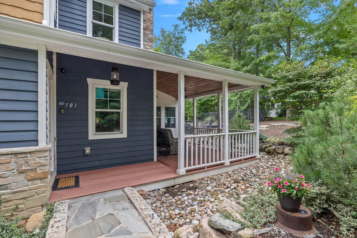 The front exterior of a house featuring blue horizontal siding and a stone facade, with a covered porch extending along the side, surrounded by landscaped rock gardens and greenery.