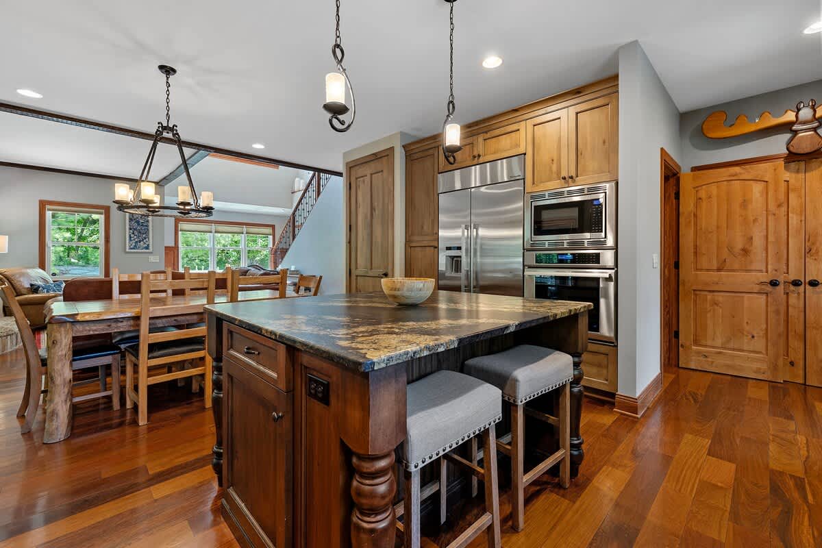 An open-concept kitchen and dining area features rich hardwood floors and wooden cabinetry, with a large island providing extra seating. Stainless steel appliances are built-in, and a staircase with a wooden railing is visible in the background, leading to an upper level.