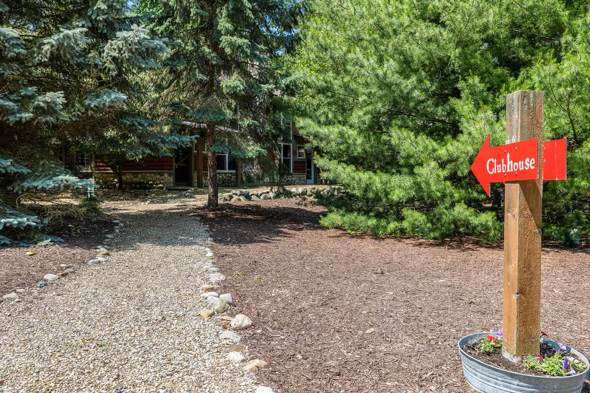 A winding gravel path lined with small rocks leads through a wooded area towards a house, with a rustic wooden sign pointing to a 'Clubhouse' on the right, surrounded by dense green foliage.