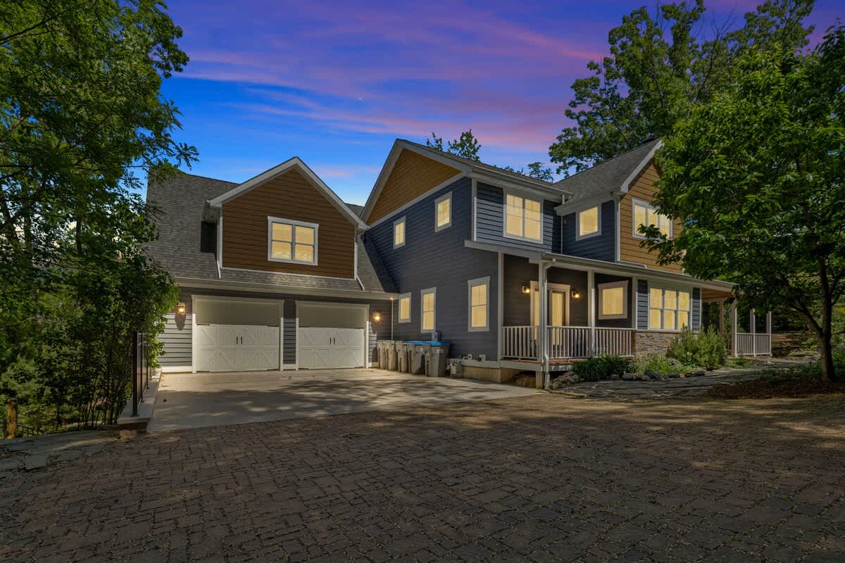 The front exterior of a large, multi-level house with blue and brown siding, illuminated by outdoor lights under a dramatic twilight sky, featuring a paved driveway and a covered porch.