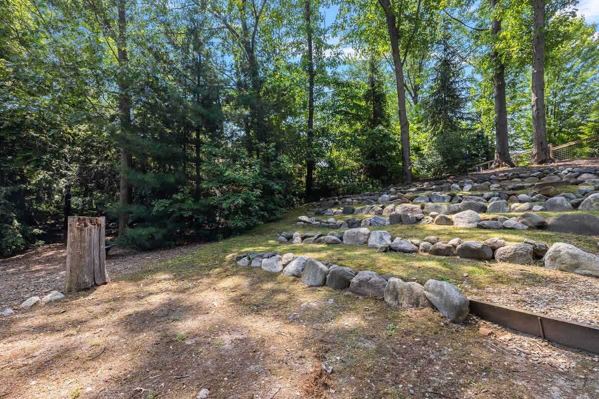 A terraced backyard area with natural rock retaining walls creating multiple levels of grassy space, surrounded by dense green trees under a bright sky, offering a natural landscape.