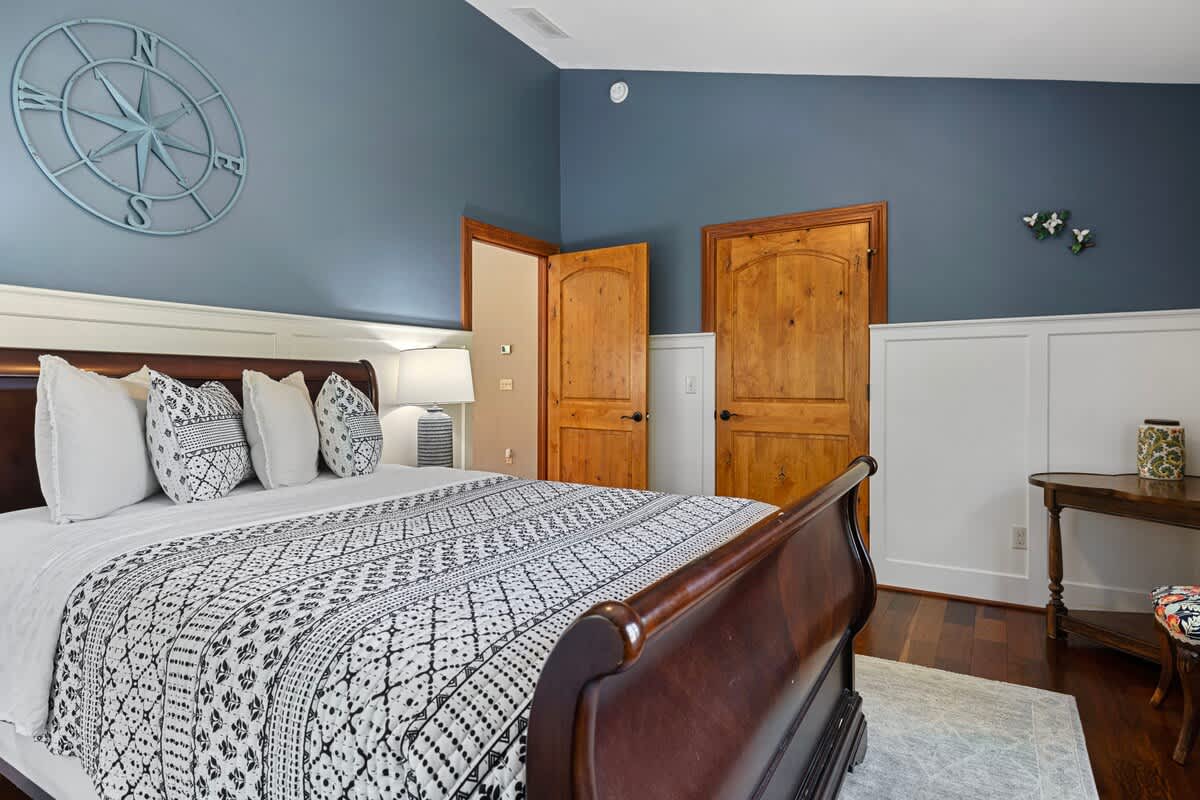 A spacious bedroom featuring a dark wood sleigh bed with white and black patterned bedding, blue walls with white wainscoting, and two wooden doors leading to other rooms. A large compass rose wall art adorns the blue wall.