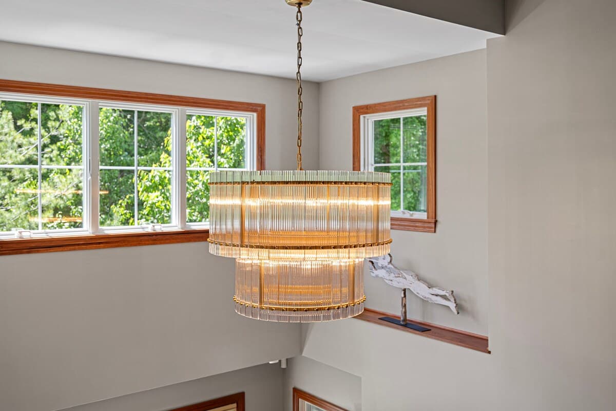 A grand tiered chandelier with glass rods hangs from a high ceiling, illuminating a staircase area with light gray walls and wooden window frames. The windows offer views of lush green trees outside.