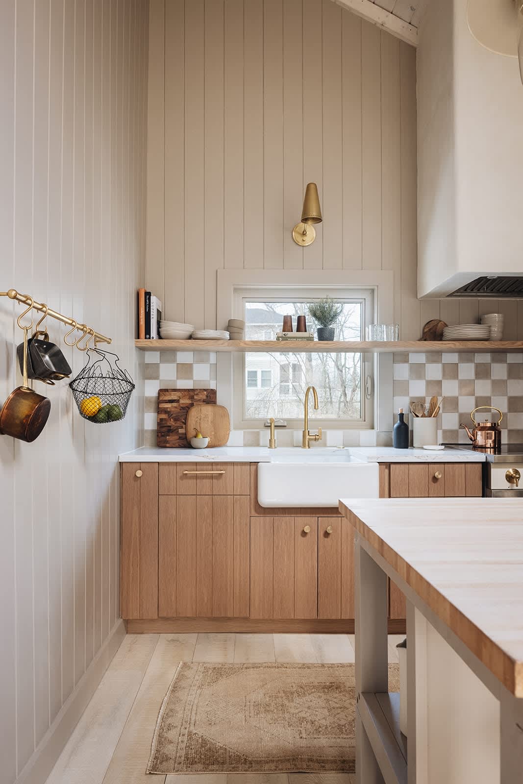 A close-up of the kitchen sink area reveals a white farmhouse sink with a brass faucet, set against a checkered tile backsplash and light wood cabinetry. Open shelving above the sink holds dishes and decor, with a brass wall sconce providing additional lighting.