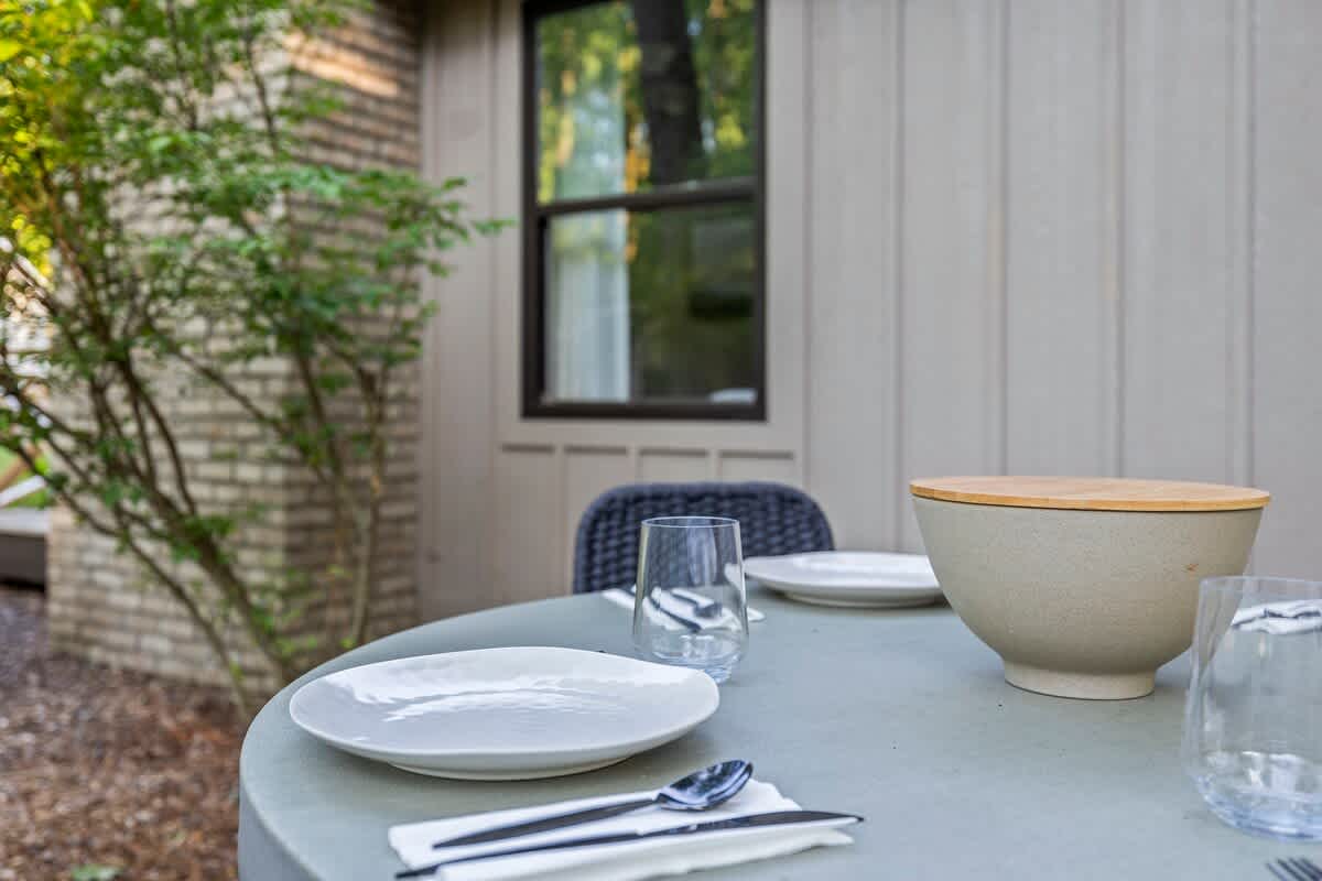 A close-up view of a modern round dining table on an outdoor deck, set with light-colored plates, clear glasses, and cutlery, alongside a large ceramic bowl with a wooden lid, against a blurred background of greenery.