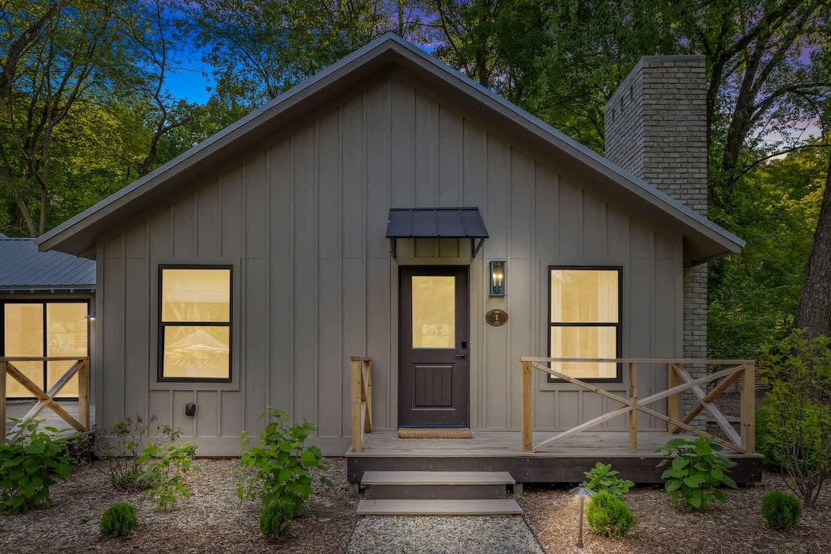 The front facade of a modern farmhouse-style cottage features light vertical siding, a dark front door with a small awning, two windows with dark frames, and a small wooden porch with railings, all set against a backdrop of trees.