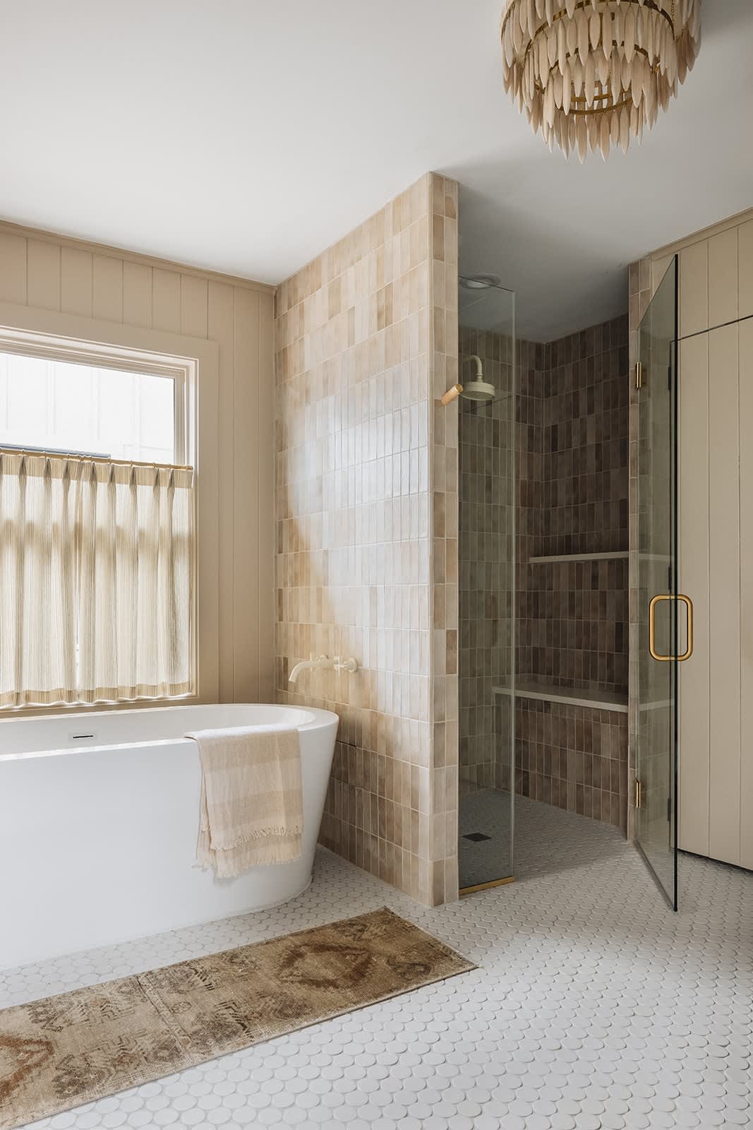 Spacious bathroom featuring a freestanding white bathtub, a walk-in shower with glass enclosure and tiled walls, and a decorative chandelier, all on white scallop-patterned floor tiles.