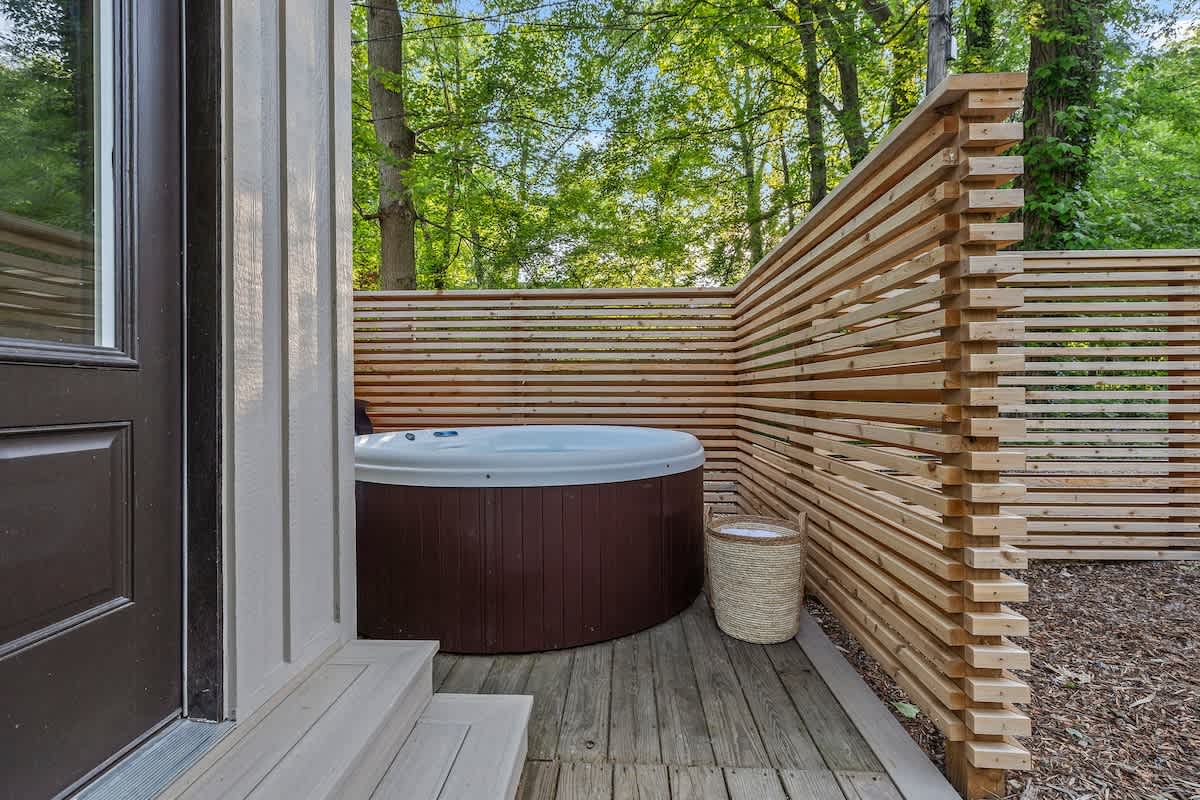 An outdoor hot tub with a dark brown base and white interior is nestled on a wooden deck, partially enclosed by a slatted wooden privacy fence. Lush green trees are visible in the background, creating a secluded ambiance.