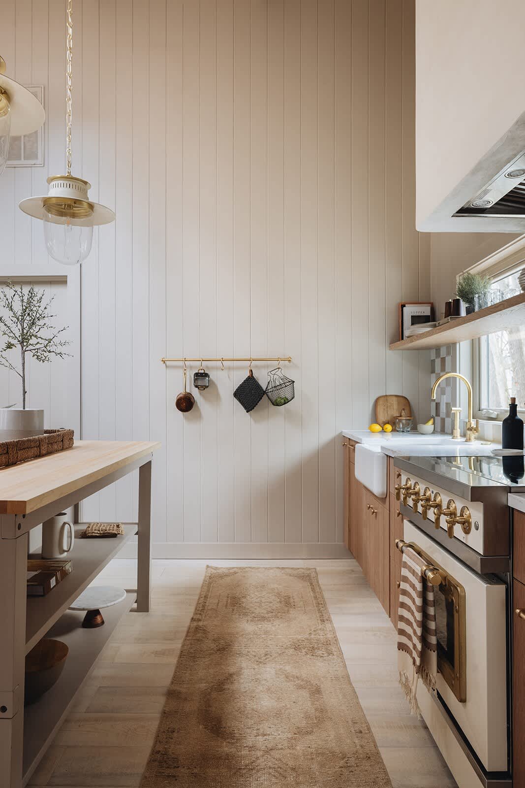 A view down the kitchen aisle shows a long runner rug on light hardwood floors, with a light wood kitchen island on the left and the main kitchen counter on the right. The counter features a white farmhouse sink, brass faucet, and a cream range with brass details, all against a checkered backsplash.